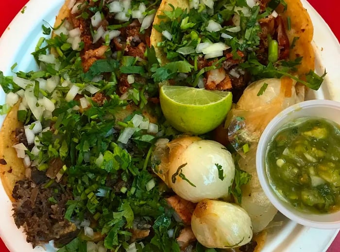 An overhead view of stacked tacos overflowing with chopped meat, diced onions, and herbs on a paper plate, accompanied by a plastic cup of salsa and a lime wedge