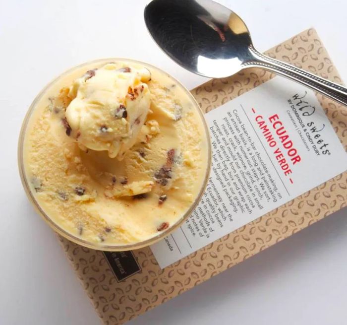 From above, a cup of ice cream filled with various mix-ins is placed atop a packaged chocolate bar, accompanied by a spoon on a white backdrop.