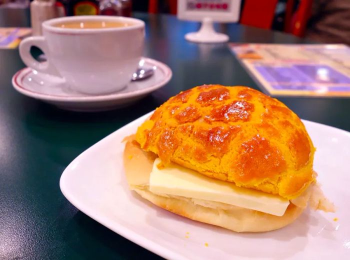 A freshly baked pineapple bun, glossy and patterned from rising in the oven, sits split open with a large slab of butter melting inside on a small plate beside a cup of coffee and a blurred menu in the background on a formica tabletop.