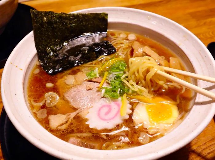 Chopsticks lift steaming ramen noodles from a bowl filled with rich broth, succulent pork, a boiled egg, nori, and fresh vegetables.