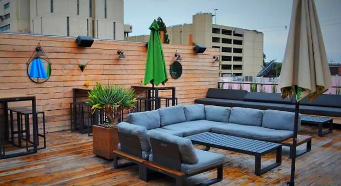 A wooden deck features U-shaped patio couches, umbrellas, potted plants, and high-top bar tables, all with a scenic city view in the background