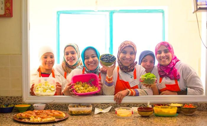 Six women lean over an interior window into the kitchen, holding bowls filled with various ingredients like olives, eggs, and greens.