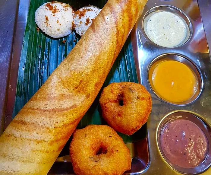 A metal tray featuring indentations for a variety of vibrant sauces sits beneath a long cylindrical dosa and two medu vadas, with a large central section covered by a banana leaf.