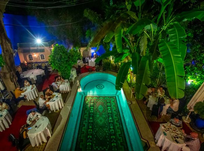 From above, a spacious pool with an ornate floor is the centerpiece of an interior courtyard/dining area, where guests are seated at tables draped in white tablecloths. Towering palm trees stretch toward the camera, and decorative structures mimic exterior architecture.