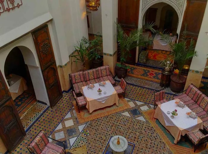 From a bird’s-eye view, a spacious open-air atrium features several tables arranged around plush couches, with tiled floors, a fountain, potted plants, and tall archways.