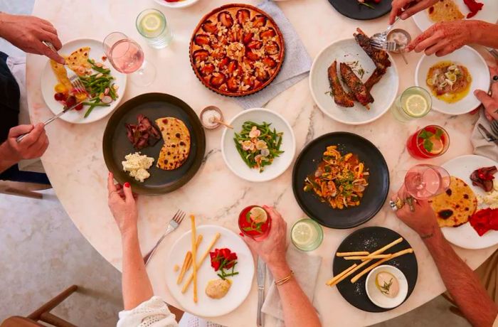 From an aerial view, a pink pastel table is filled with diners enjoying a variety of dishes, including salads, tarts, ribs, and flatbreads.