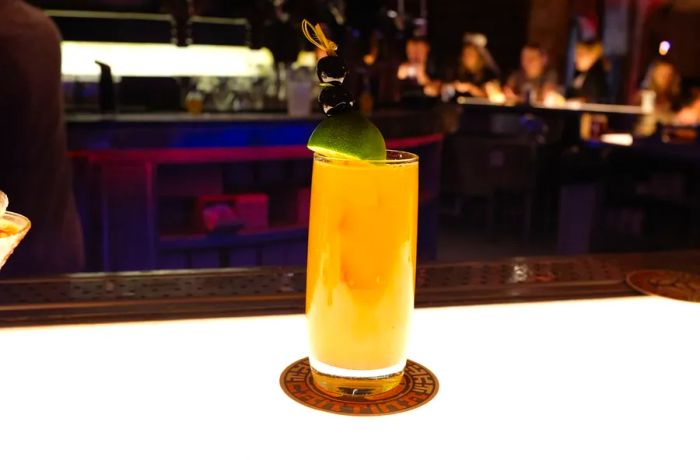 A highball glass filled with an orange cocktail garnished with a lime wedge, resting on a coaster at a brightly lit bar, with blurred customers in the background.