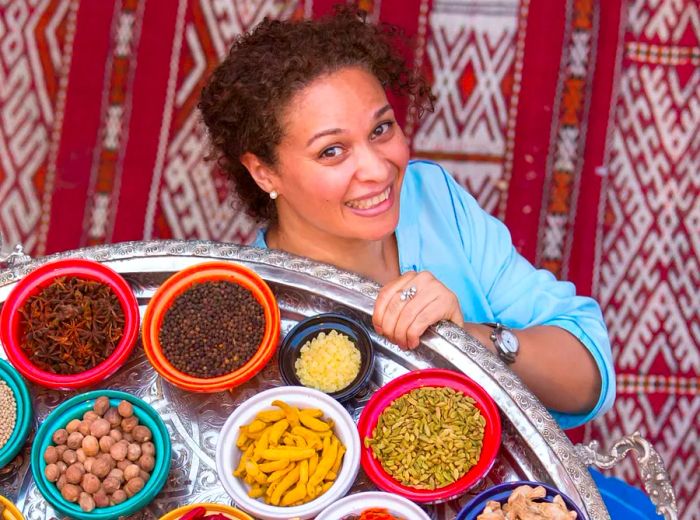 From an elevated view, a woman stands on a decorative carpet, holding a large silver platter filled with an array of small, colorful bowls containing fresh ingredients.