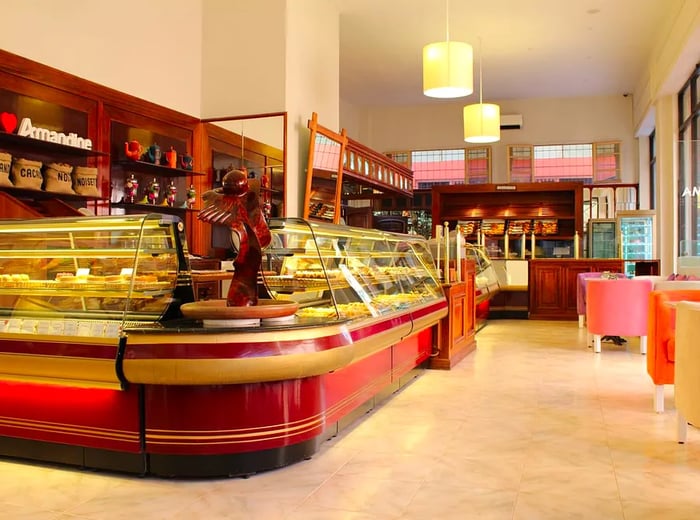 A long, elegantly curved pastry display inside a bright patisserie, featuring pendant lights, marble floors, and wooden shelves showcasing products and branding.