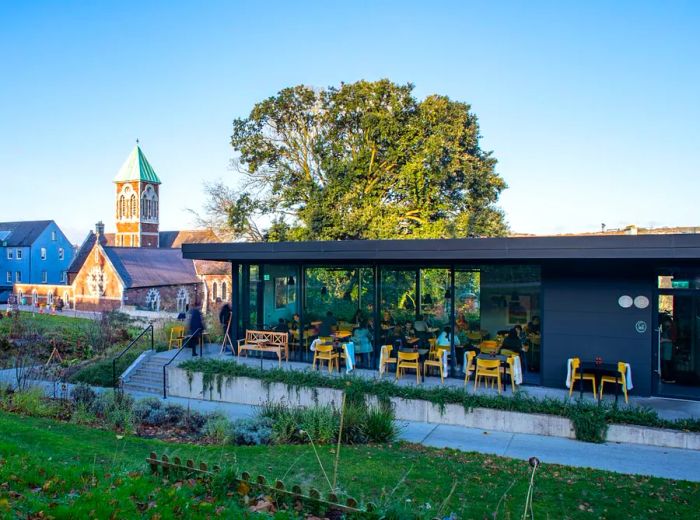 A sleek, modern building with expansive floor-to-ceiling windows and a dark exterior overlooks lush greenery, with an 18th-century church visible in the background.