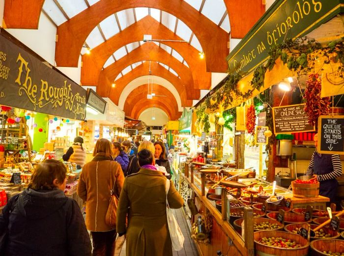 A series of market stalls stretch away into a lofty covered atrium, with customers strolling along the aisles. The nearest stand showcases barrels of olives, complemented by handwritten chalk signs promoting various products.