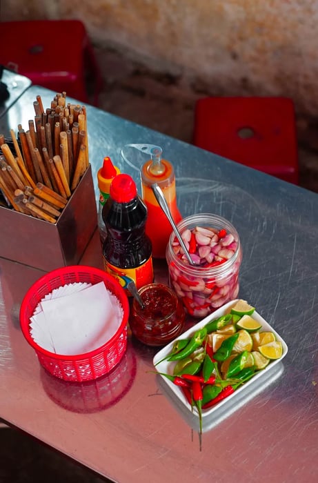 A jar of chile sauce rests atop a metal table, accompanied by fresh chiles, lemon wedges, pickled garlic, and various bottled sauces.