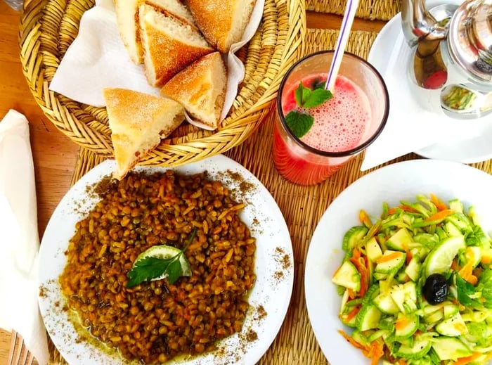 Aerial view of a rustic wooden table displaying a plate of grains, a vegetable platter, and a basket of triangular flatbreads, alongside a tall glass of fruit juice garnished with fresh mint.