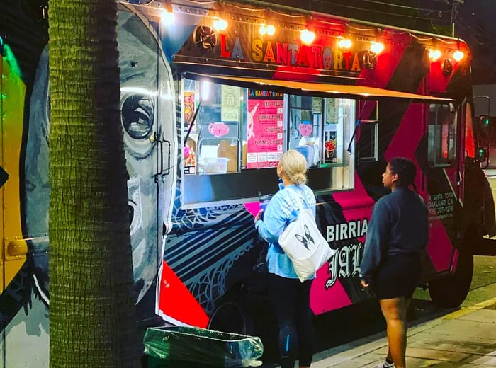 Two women stand by a brightly lit food truck adorned with a masked luchador design