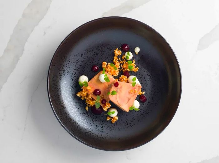 An aerial view of a dark plate featuring two pieces of foie gras adorned with dots of gel, a crumbly topping, and finely chopped herbs.