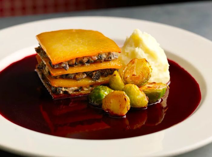 A beautifully layered galette with ingredients spilling out sits alongside a generous scoop of mashed potatoes and a serving of chopped Brussels sprouts, all drizzled with dark gravy on a white plate.