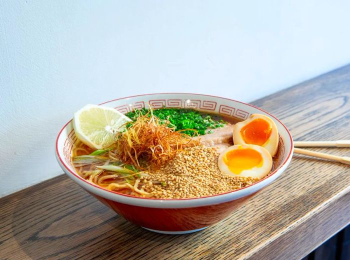 A bowl of ramen sits on a wooden counter against a neutral wall. Floating in the savory broth are a boiled egg, diced greens, fried toppings, and noodles.