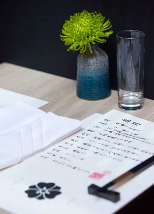 A Japanese menu adorned with a delicate floral illustration rests on a light wooden table, illuminated by a pendant light. Nearby, chopsticks sit on a holder beside a folded napkin and a petite vase.