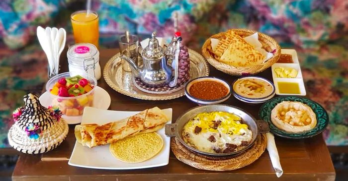 A quaint wooden table showcases an array of dishes, including flatbreads, a fruit bowl, various dips, a tray of tea, and a glass of orange juice.