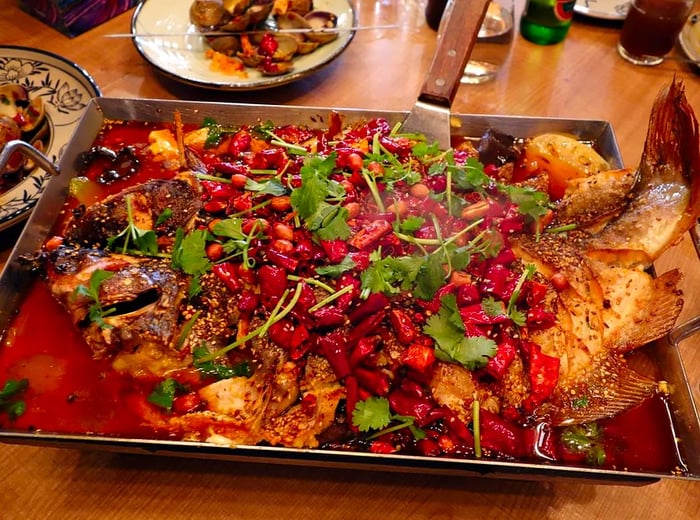 A metal tray holds a whole roasted fish topped with a vibrant mix of chiles, herbs, peanuts, and seeds, surrounded by various other dishes on the table.