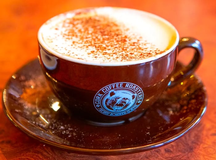 A frothy cappuccino, sprinkled with spices, is served in a wide-brimmed branded mug from Cork Coffee Roasters, resting on a saucer.