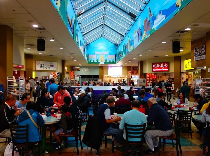 In a bright atrium with a large skylight, patrons gather at tables and stroll among various vendors lining the outer edges, creating a lively atmosphere.