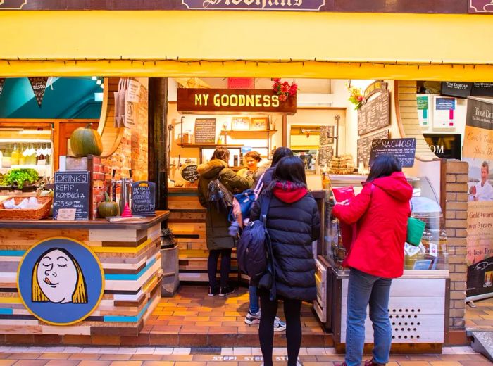 Shoppers queue at a vibrant market stall adorned with a sign that reads My Goodness, featuring a whimsical illustration of a woman enjoying a treat on its exterior.