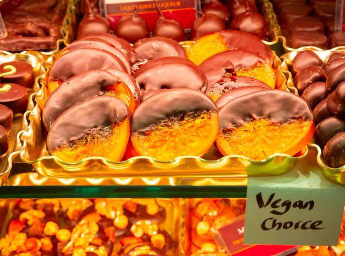 A tray of chocolate-dipped orange slices sits in a deli case, surrounded by an assortment of other chocolate treats. A post-it note labeled 'vegan choice' is affixed to the edge of the tray.