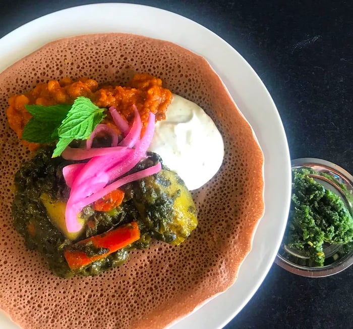 A bird’s-eye view of a plate nearly overflowing with soft injera flatbread, adorned with vibrant curries and yogurt, accompanied by a small bowl of green dip.