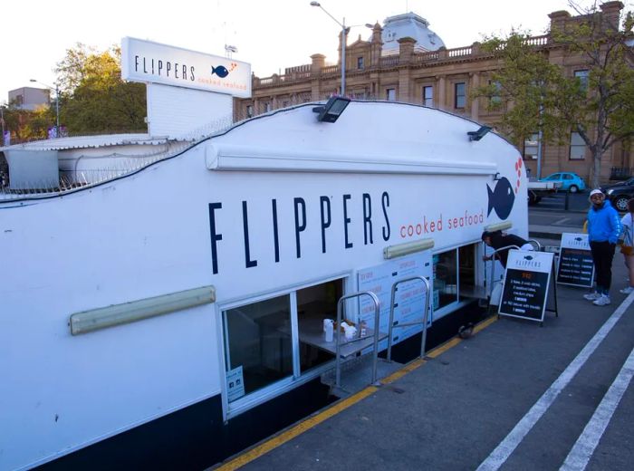 A floating eatery anchored at a pier, featuring service windows positioned just above the water's edge, with the name Flippers displayed prominently on the side of the boat.