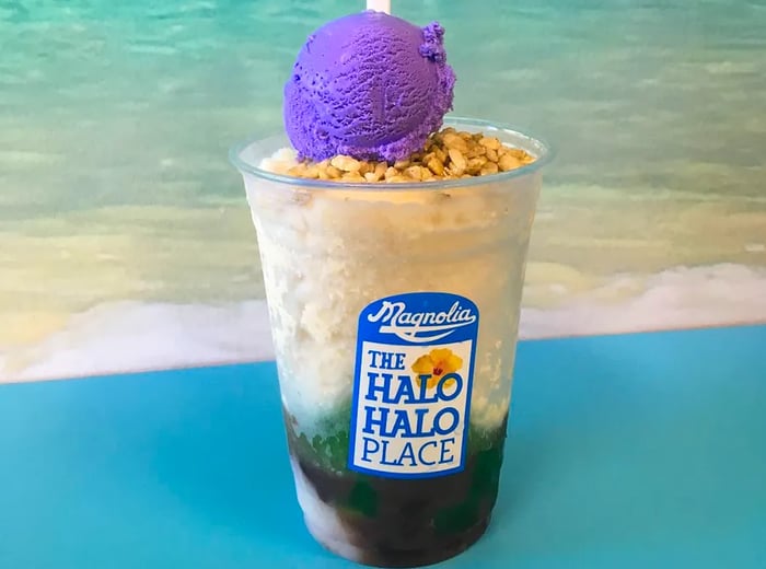 A plastic cup layered with colorful flavored ice, fruits, and nuts, topped with a scoop of vibrant purple ube ice cream skewered with a popsicle stick against a backdrop of the ocean.