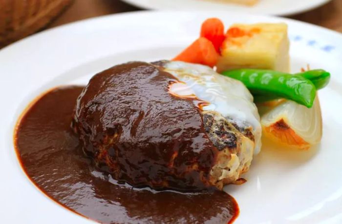 A juicy hamburg steak patty drizzled with various sauces that pool around it on a white plate, accompanied by slices of cooked vegetables in the background.