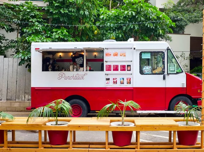 A view of a food truck from inside a cafe, featuring a long table and seating arranged by a large window.