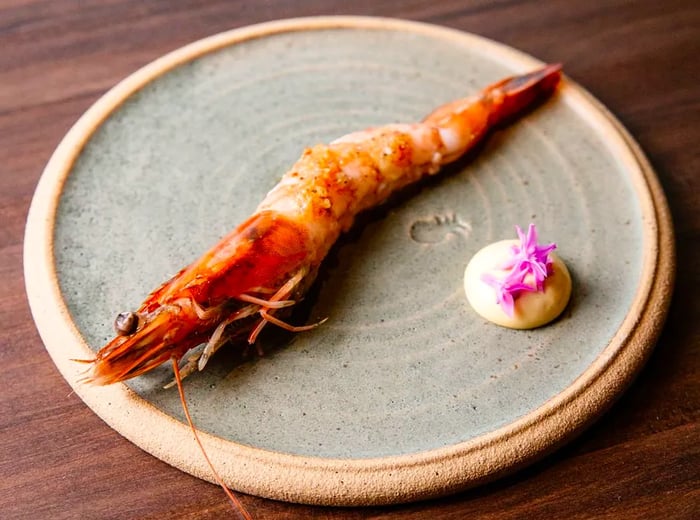 A single large shrimp on a plate, accompanied by a vibrant sauce and garnished with a flower.