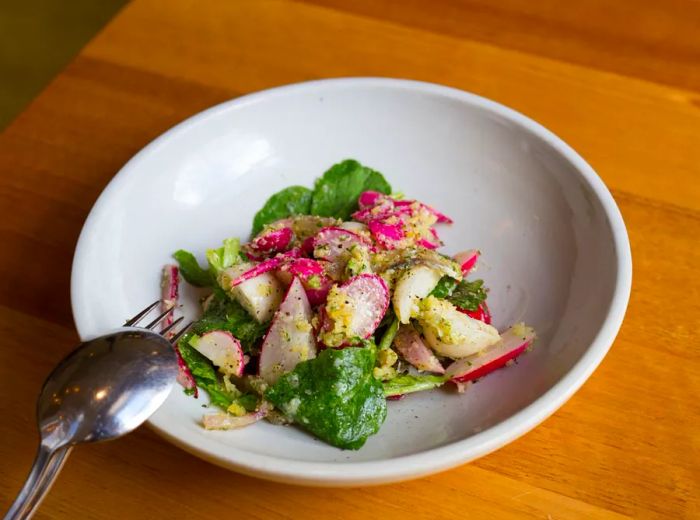 A shallow bowl filled with chunks of radish mixed with greens, pieces of salmon, and cheese, accompanied by a spoon and fork resting on the rim, all set on a wooden tabletop.