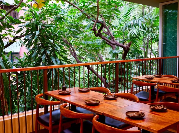 A wooden six-top table set with small plates, nestled beside a railing with thick trees in the background.