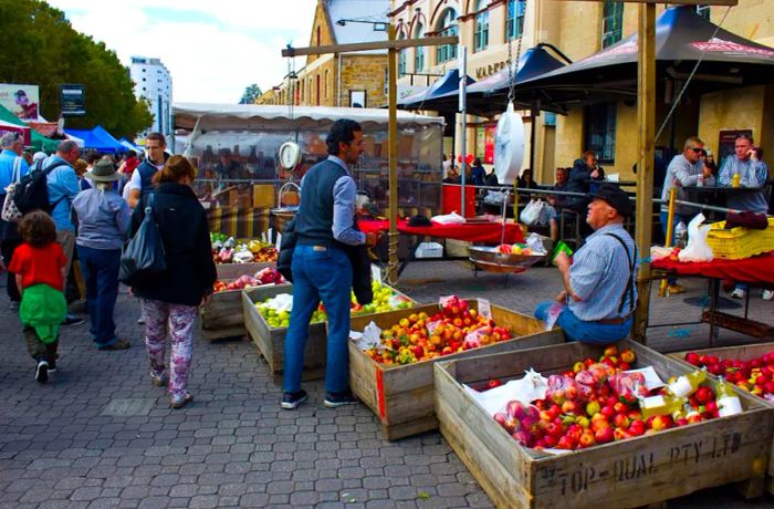 A vendor at a farmers market sits among crates filled with apples, chatting with a customer, as passersby stroll by on the left and others enjoy their meals at tables on the right.