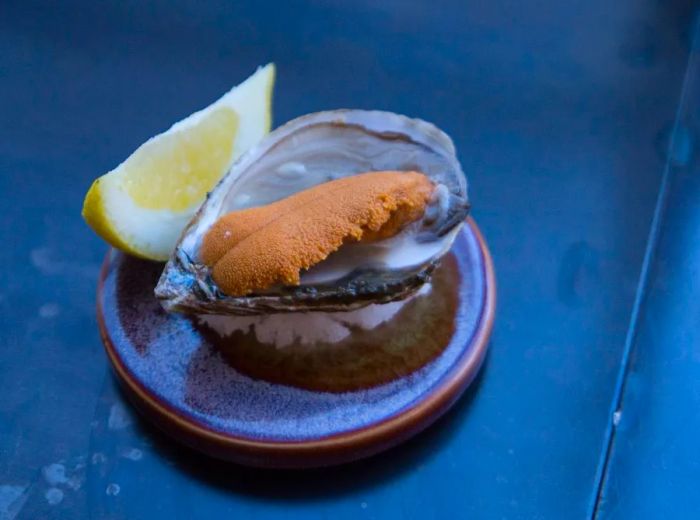 A freshly shucked oyster, lavishly topped with a generous dollop of sea urchin roe, rests on a small plate accompanied by a lemon wedge on a cool, dark surface.