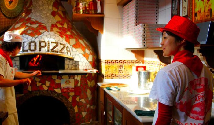 A chef retrieves a pizza from a blazing pizza oven, flames visible inside, while a server observes, with a prep station and stacked pizza boxes in the background.