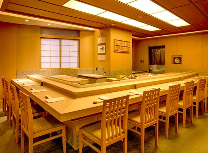 An empty L-shaped sushi counter flanked by simple chairs in a dimly lit, paneled room, featuring a screen door on one wall.