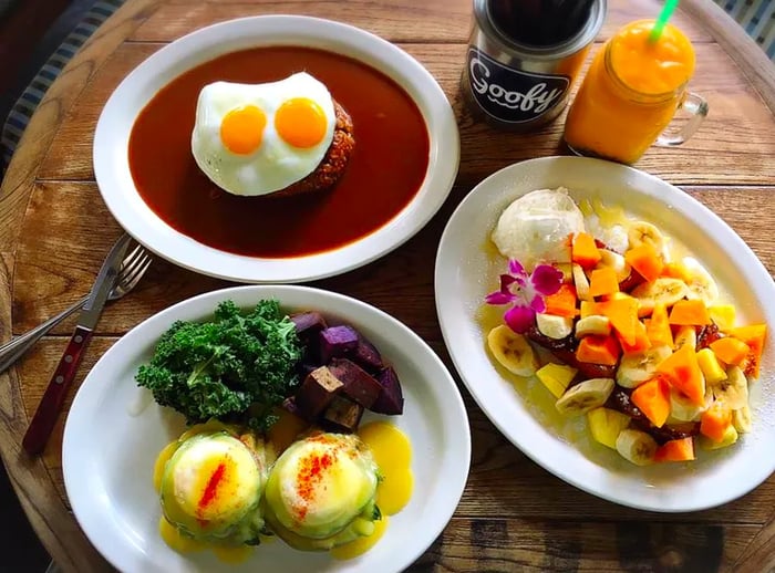 A top-down view features a barrel-shaped table adorned with eggs benedict alongside salad, fruit-covered French toast, and loco moco.