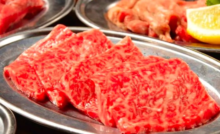 An arrangement of six to seven slices of raw meat on a metal platter, with other meat dishes softly blurred in the background.