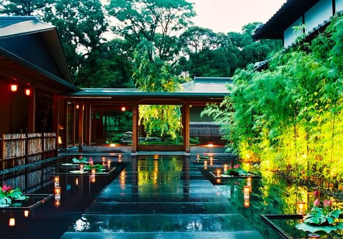 The courtyard of a Tokugawa residence, featuring shallow pools adorned with plants and lanterns, tall greenery on one side, and large trees in the background, with a covered walkway connecting two buildings.