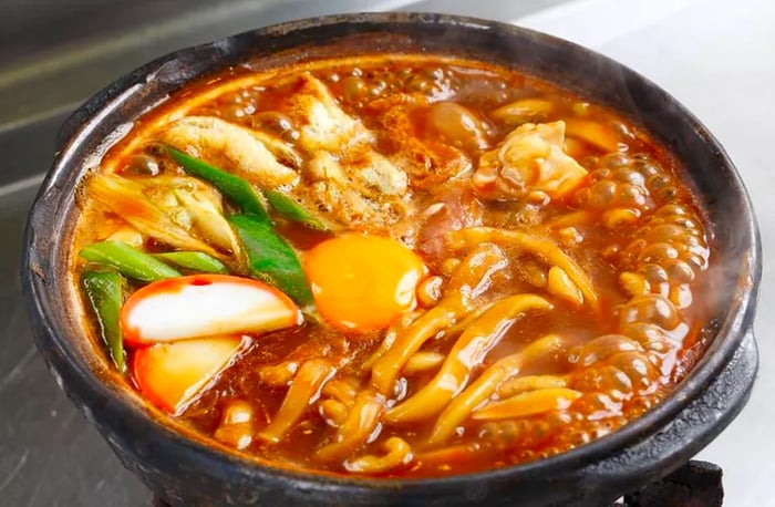 A steaming bowl of noodle soup served in a clay pot, adorned with egg yolk, slices of radish, fresh greens, and a generous portion of noodles on top.