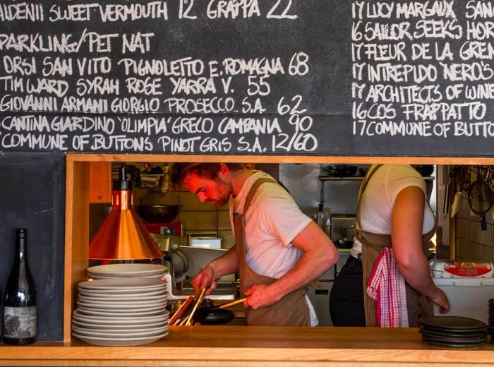 A large slate wall displays a chalk menu in the top half, while the bottom features a long cut-out window leading to the kitchen, where cooks can be seen at work, along with a shelf showcasing plates and bottles of wine.