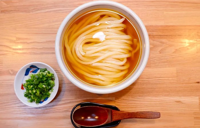 Aerial view of a light wooden table featuring a white bowl filled with delicate udon noodles in broth, alongside a spoon resting in its holder, and a bowl of greens for garnish.