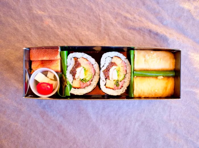 A bird’s-eye view showcases a long, segmented box filled with two sizable cut maki rolls and an array of other dishes.