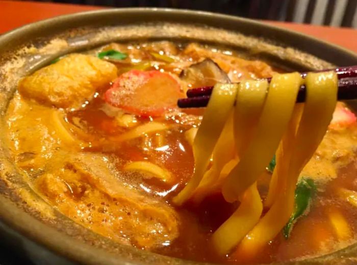 Chopsticks lift noodles from a steaming clay pot filled with broth, tender meat, tofu, and a medley of vegetables.