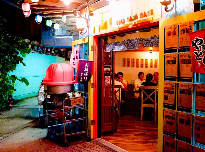 The entrance to the restaurant is adorned with string lights and Japanese signage, leading into a turquoise alleyway on one side.