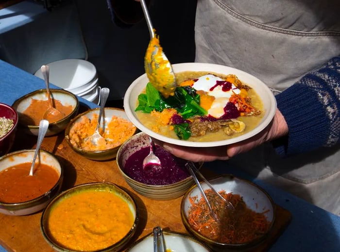 A server stands over a prep station filled with eight bowls of various ingredients, cradling a bowl of congee in one hand while using the other to ladle vibrant greens atop colorful toppings.